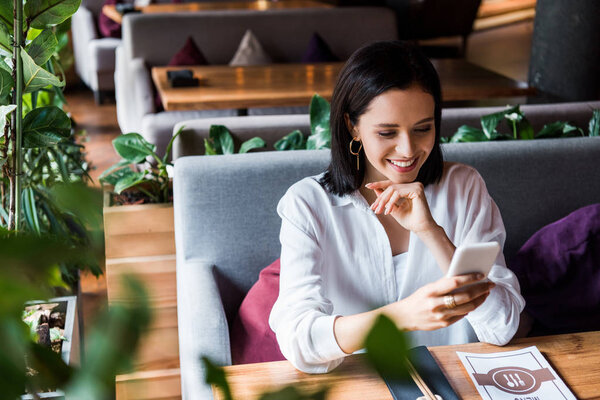 selective focus of cheerful woman sitting in restaurant and using smartphone 