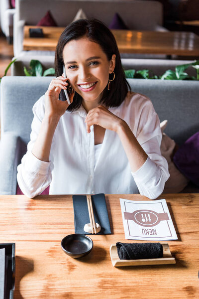 happy woman talking on smartphone near menu on table 