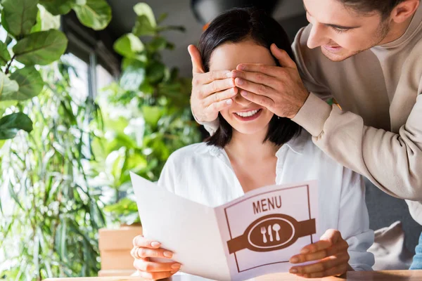 Cheerful man closing eyes of cheerful woman holding menu in restaurant — Stock Image Cheerful Man Closing Eyes Cheerful Woman Holding Menu Restaurant Stock Photo