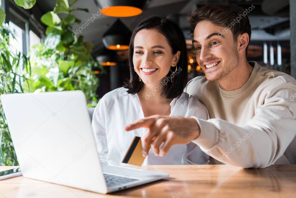 selective focus of happy woman looking at laptop man holding credit card and pointing with finger 