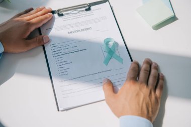 cropped view of doctor sitting at table with blue awareness ribbon on medical report clipart