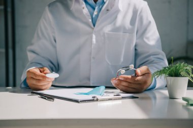 Masada oturan ve steteskopu mavi farkındalık kurdelesinin yanında tutan doktorların kesikli görüntüsü.