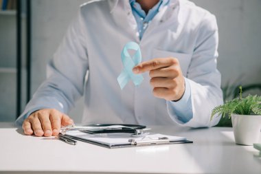 Doktor masasında oturup mavi farkındalık kurdelesi gösteriyor.