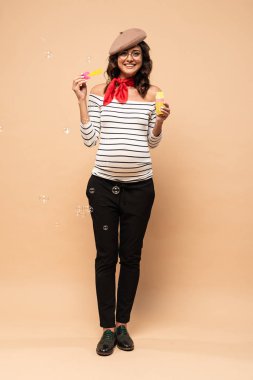 pregnant french woman in beret holding bottle with soap bubbles on beige background  clipart