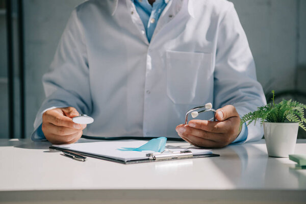 обрезанный вид доктора, сидящего за столом и держащего стетоскоп возле синей ленты осознания
