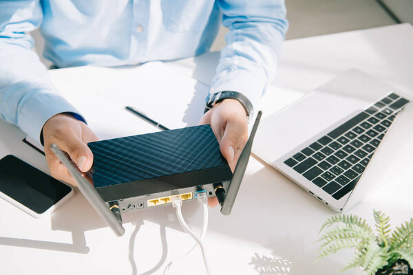 partial view of businessman holding plugged router near smartphone and laptop