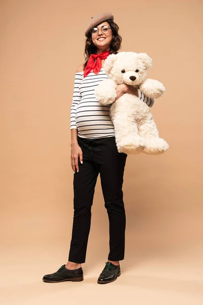 身穿贝雷帽的法国孕妇笑着把泰迪熊抱在米色背景上 — 图库照片