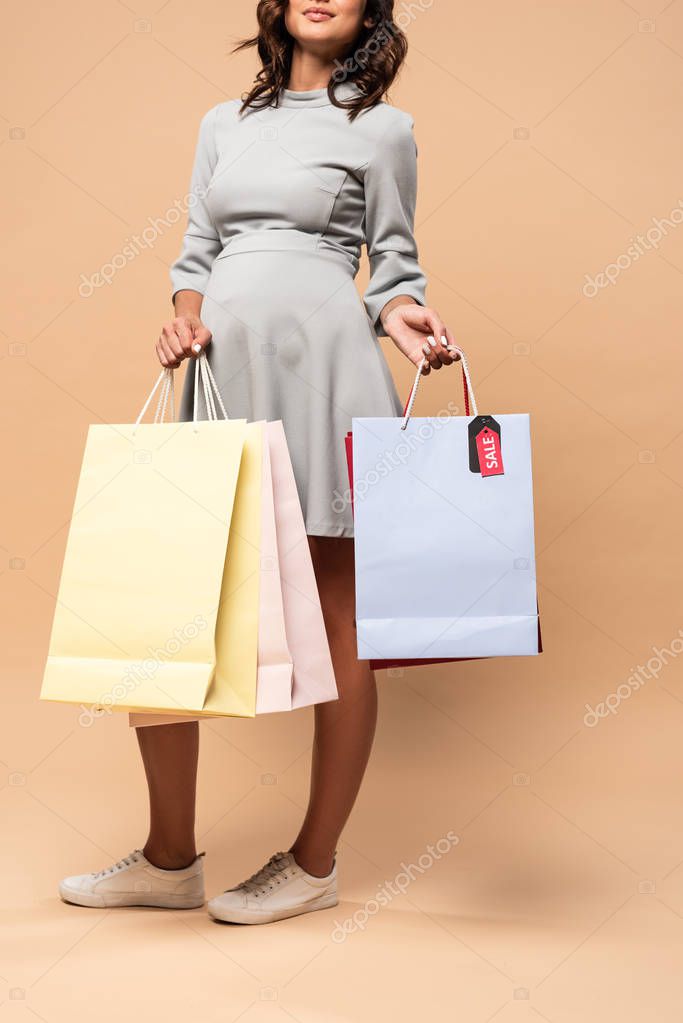 Cropped view of pregnant woman in grey dress holding shopping bags on beige background