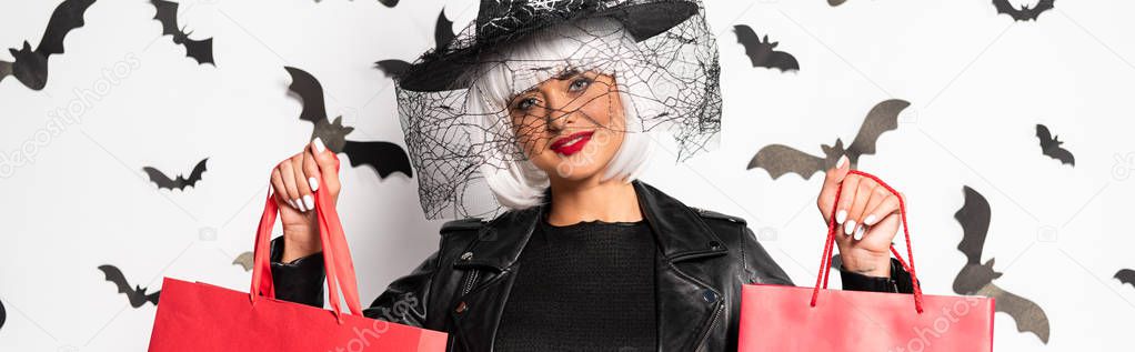 panoramic shot of attractive woman in witch hat and wig holding shopping bags in Halloween