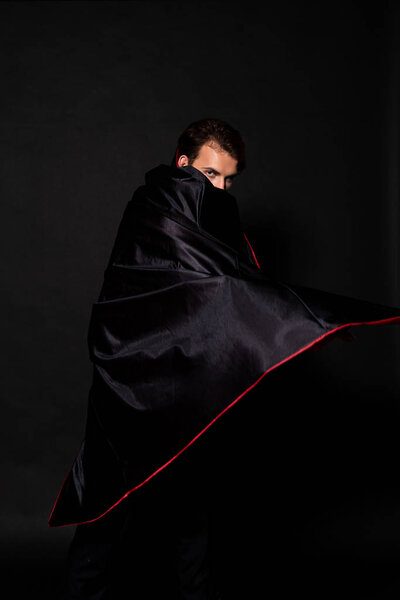 man covering face with cloak of halloween costume on black 