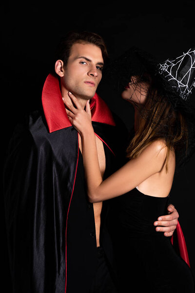 woman in witch hat touching neck of man in halloween costume isolated on black 