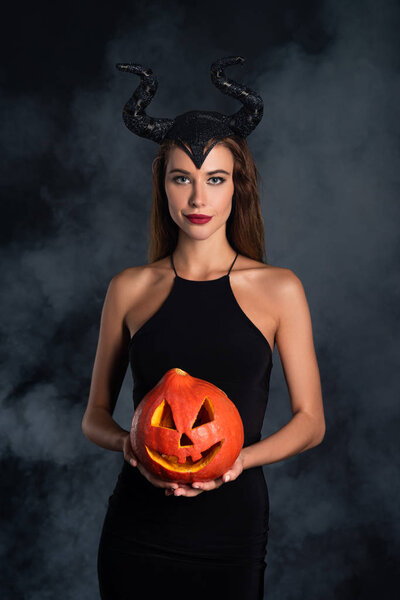attractive woman with horns holding halloween pumpkin on black with smoke 