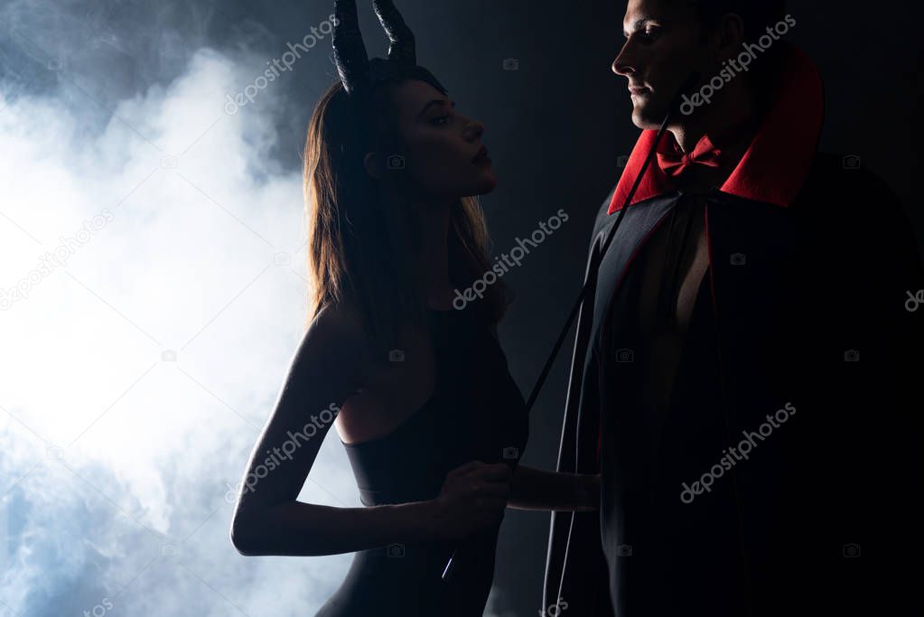 handsome man in cloak looking at girl with horns holding flogging whip on black with smoke 