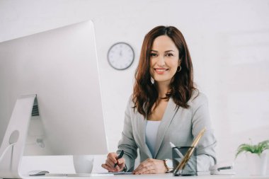 beautiful, smiling secretary looking at camera while sitting at workplace near computer monitor clipart