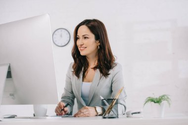 attractive, smiling secretary looking at computer monitor and writing in notebook clipart