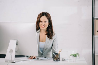 attractive, smiling secretary looking at camera while sitting at workplace clipart