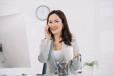 Neşeli sekreter kahve fincanı tutuyor ve iş yerinde otururken akıllı telefonla konuşuyor.