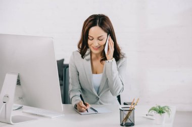 smiling secretary talking on smartphone and writing in notebook while sitting at workplace clipart