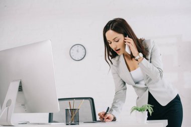 attentive secretary talking on smartphone and writing in notebook while standing at workplace clipart