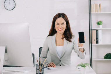 neşeli sekreter iş yerinde otururken boş ekranlı akıllı telefonu gösteriyor