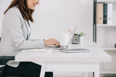 cropped view of smiling secretary using laptop while sitting at workplace in office clipart