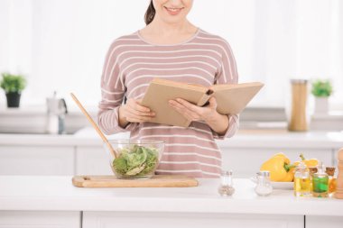 Mutfak masasının yanında sebze salatasıyla dururken gülümseyen bir kadının fiş okuduğu görüntüler.