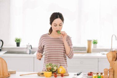 pretty, young woman tasting salad while standing at kitchen table with fresh vegetables