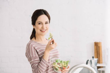 Genç, gülümseyen bir kadın sebze salatası yerken kameraya bakıyor.