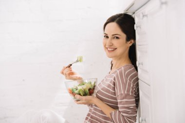 Mutfakta oturmuş sebze salatası yerken kameraya bakan mutlu genç bir kadın.