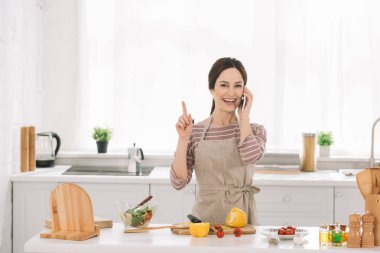 Önlüklü neşeli kadın mutfak masasının yanında sebzelerle durup akıllı telefondan konuşurken fikir işareti gösteriyor.