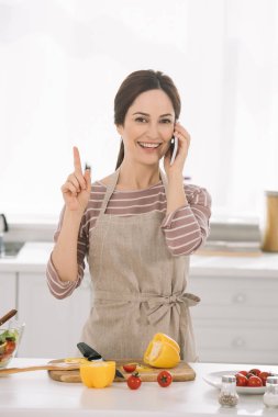 Önlüklü neşeli kadın mutfak masasının yanında sebzelerle durup akıllı telefondan konuşurken fikir işareti gösteriyor.