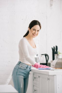 smiling housewife looking at camera while wiping kitchen table with rag