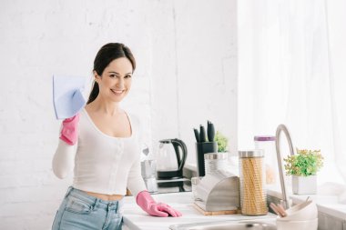 happy housewife looking at camera while standing in kitchen and holding rag