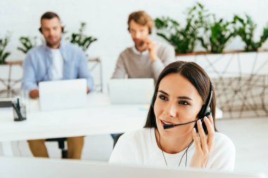 selective focus of broker touching headset near coworkers in office  clipart