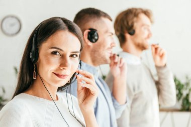 selective focus of attractive broker touching headset near men in office  clipart