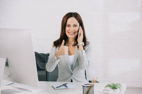 улыбающаяся секретарша показывает смартфон с чистым экраном и большим пальцем, сидя на рабочем месте
