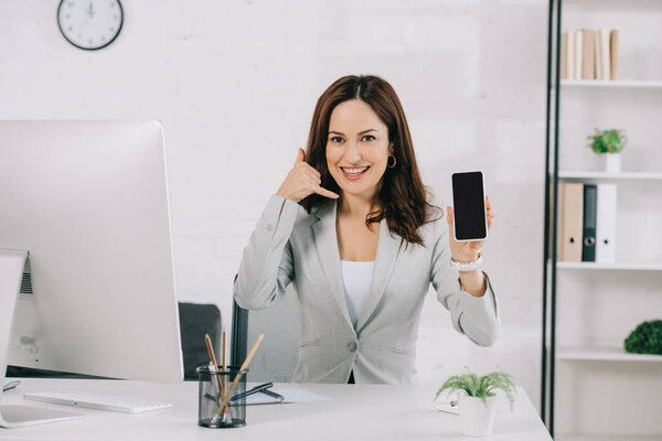 smiling secretary showing smartphone with blank screen and lets drink gesture while sitting at workplace