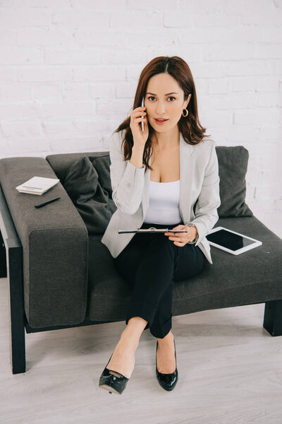 attentive young secretary talking on smartphone while sitting on couch near digital tablet