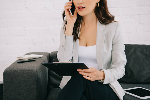 cropped view of young secretary holding clipboard and talking on smartphone in office