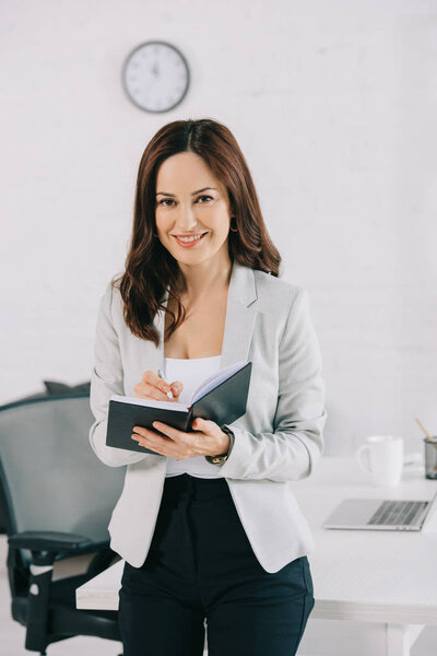 beautiful, smiling secretary looking at camera while writing in notebook