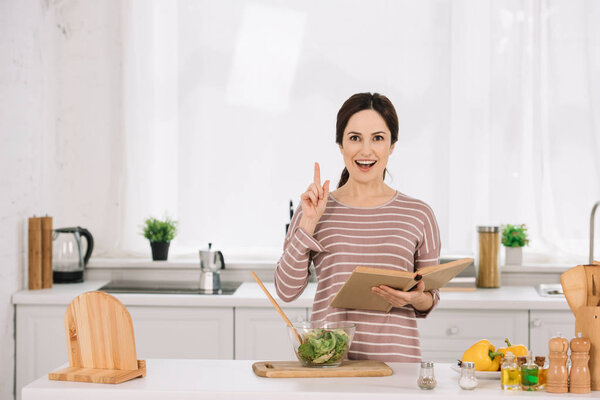 attractive, smiling woman showing idea sign while standing at table with fresh vegetables and holding receipt book