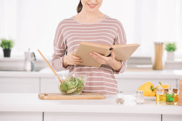 cropped view of smiling woman reading receipt book while standing at kitchen table near bowl with vegetable salad