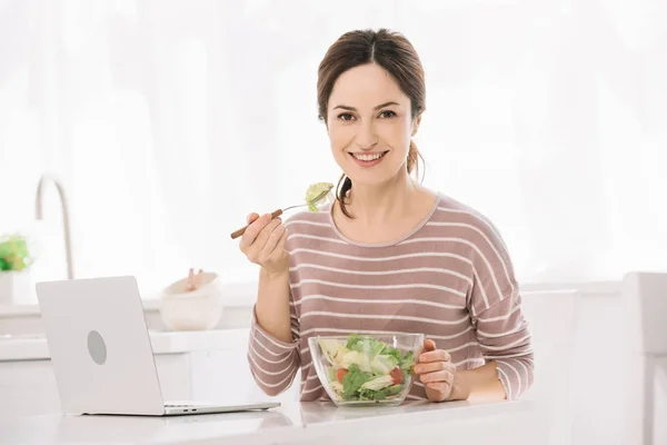 ラップトップの近くのテーブルに座って野菜サラダを食べながらカメラを見ている若い幸せな女性 — ストック写真