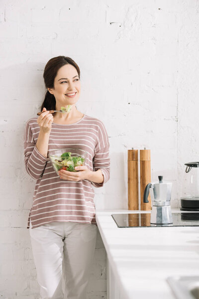 young, happy woman looking away while standing near wall and eating vegetable salad