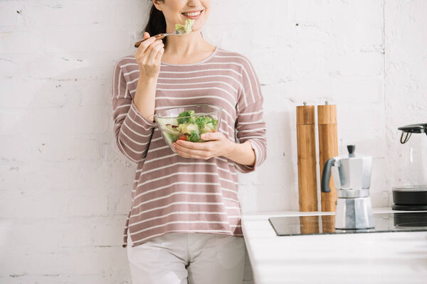 cropped view of smiling woman standing near wall and eating vegetable salad