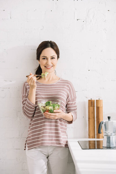 young, happy woman looking at camera while standing near wall and eating vegetable salad