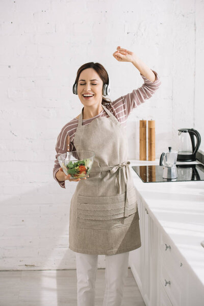happy young woman in headphones dancing and singing while holding bowl with salad