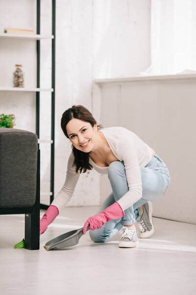 cheerful housewife looking at camera while sweeping floor with brush and scoop