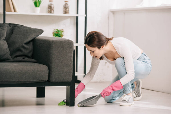 young housewife sweeping floor near sofa with brush and scoop