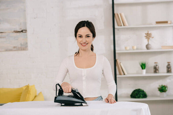 cheerful housewife smiling at camera while ironing on ironing board